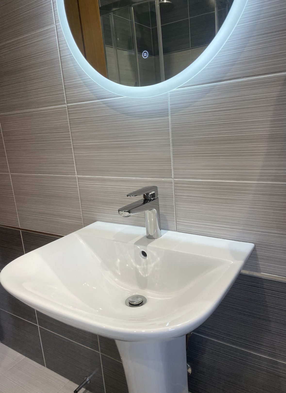 New white pedestal basin with a chrome mixer tap beneath a round illuminated mirror on grey tiles