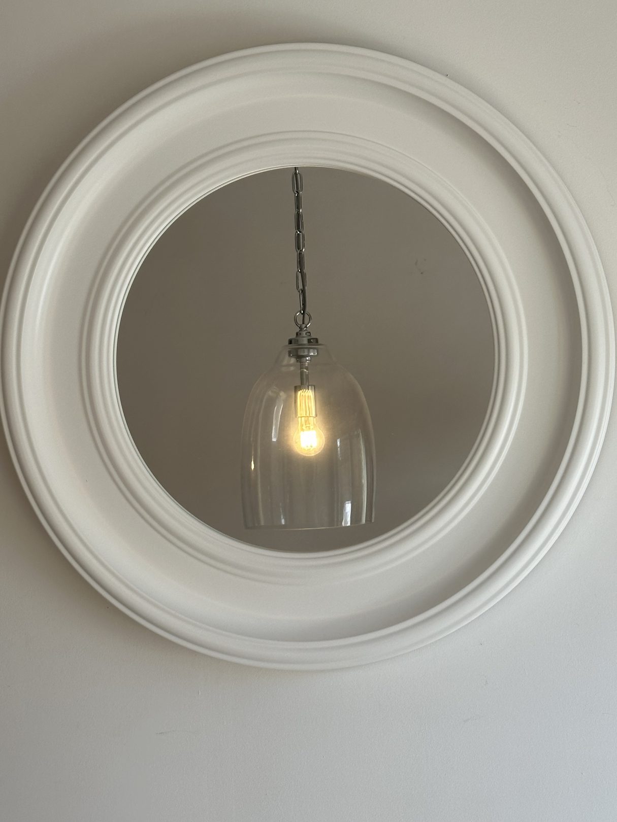 Large round white-framed mirror hung on a wall, reflecting a glass pendant light